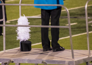 High school marching band conductor stands on metal lift with shako hat with plumes resting near feet during football game